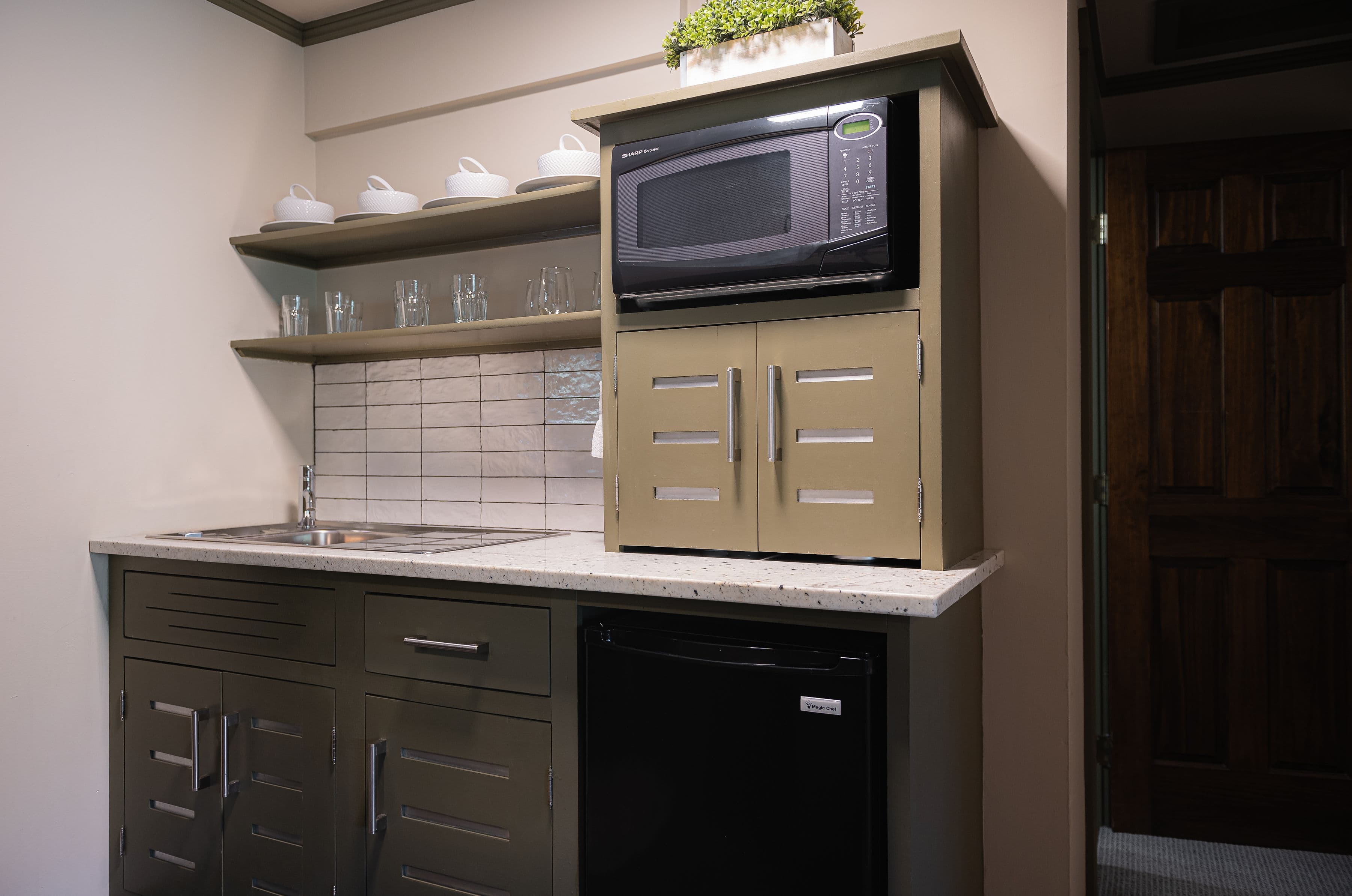 kitchenette with mini-fridge, microwave, cabinet with coffee maker, shelves with dishes and metal sink.