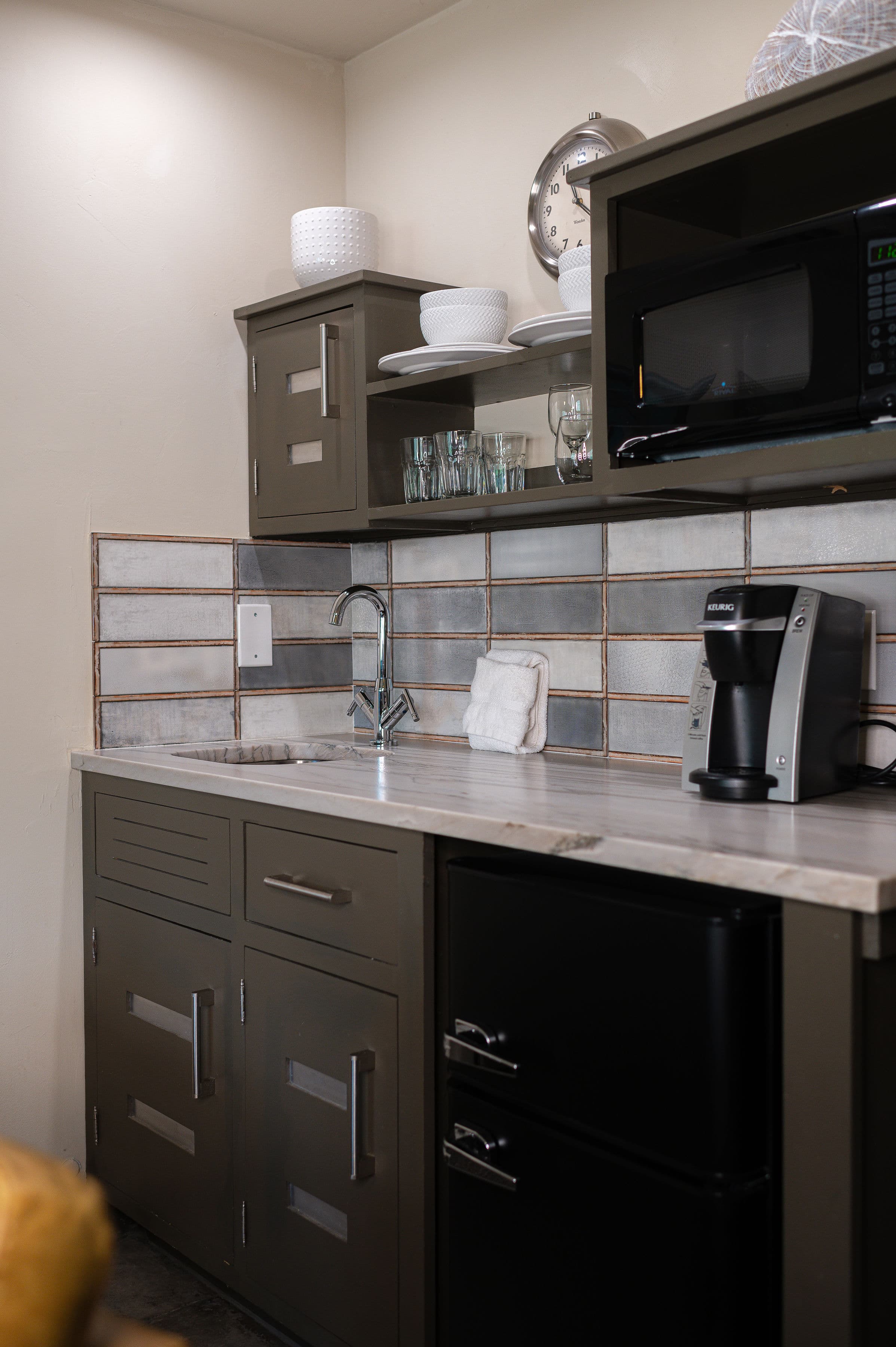 kitchenette with mini-fridge, microwave, cabinet with coffee maker, shelves with dishes and metal sink.