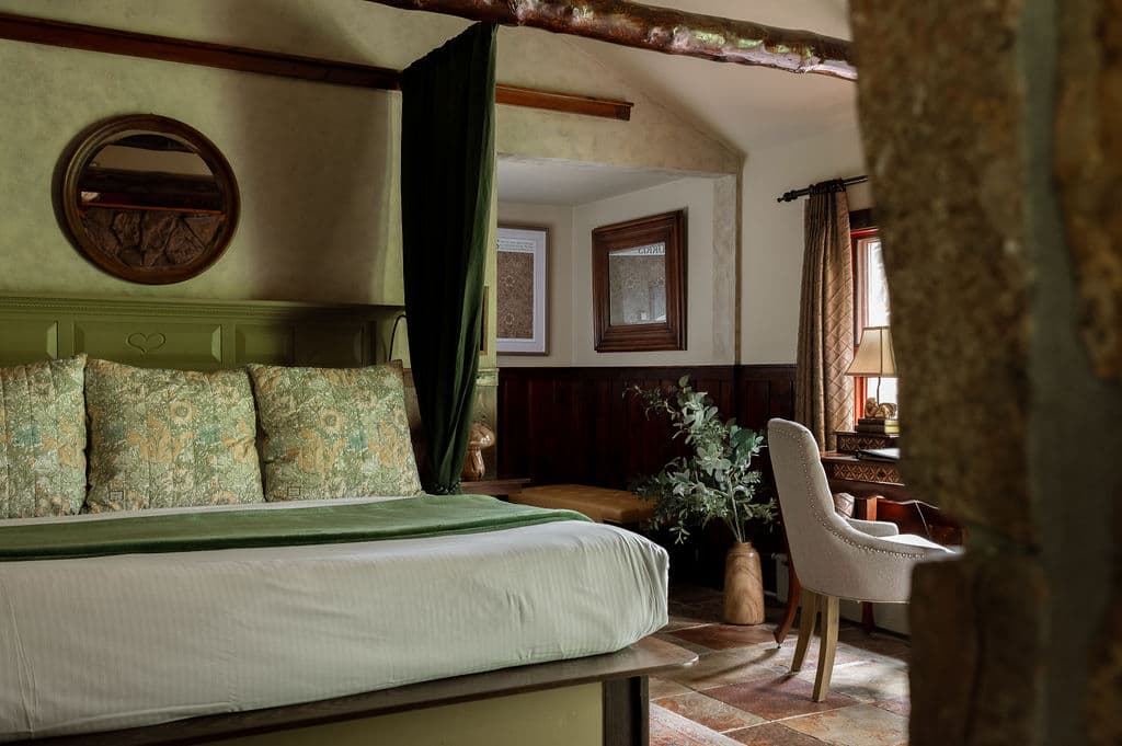 King bed with soft brown throw, Aztec decorative pillows and log beams over the bed. moss rock Gas fireplace at foot of bed