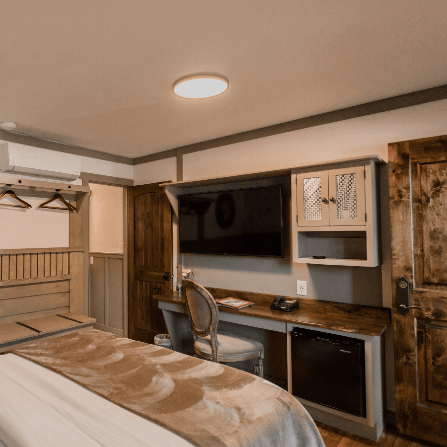 natural edge wood desk at foot of bed with plush sitting chair, mounted flat screen TV, mini fridge, and cabinet with coffee maker.