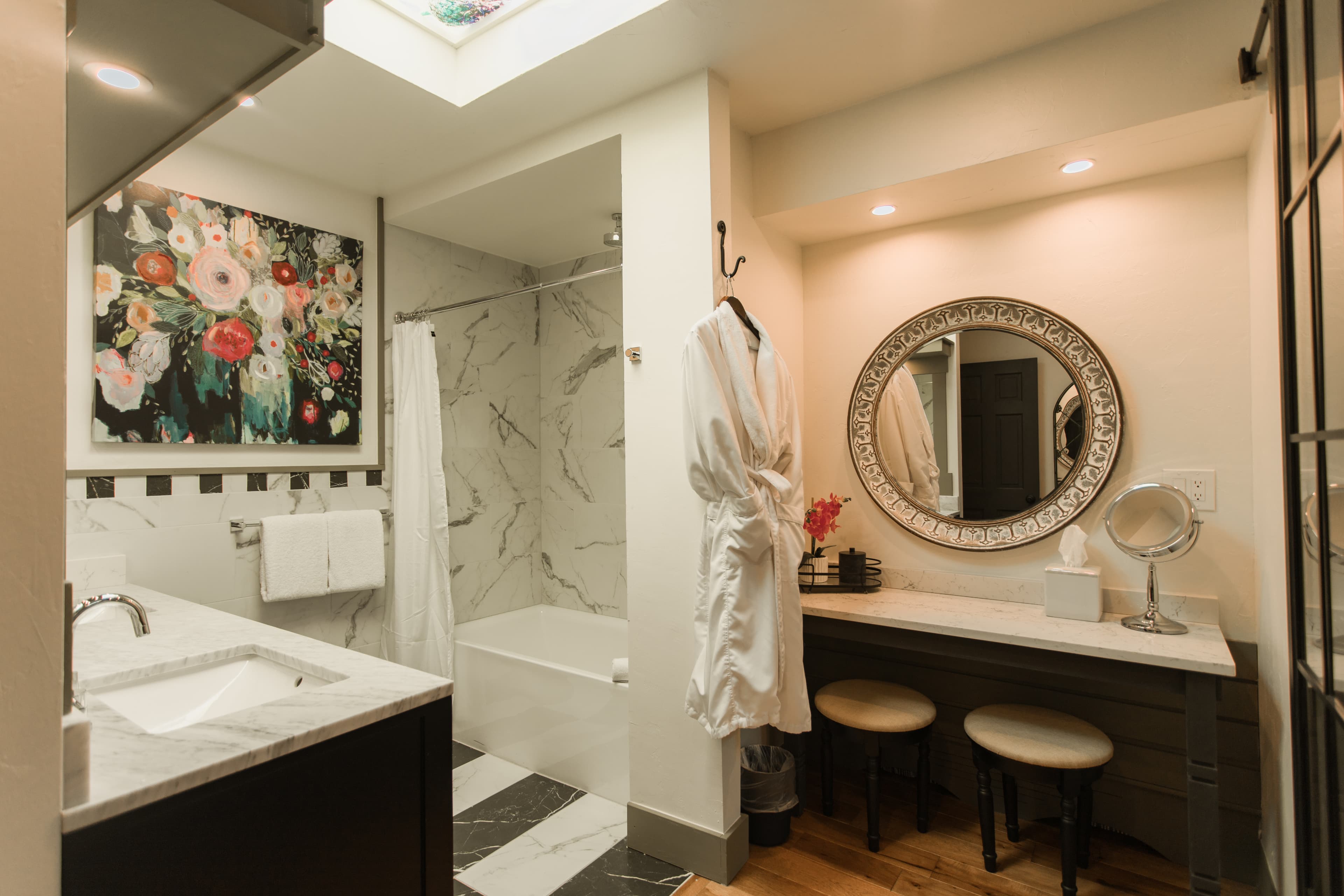 A modern bathroom with marble accents, a floral painting, and a vanity area featuring a round mirror and two stools.