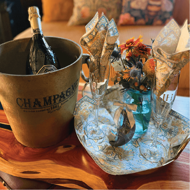 A champagne bottle in a metallic ice bucket next to two glasses, a small gift, and a floral arrangement on a decorative tray.