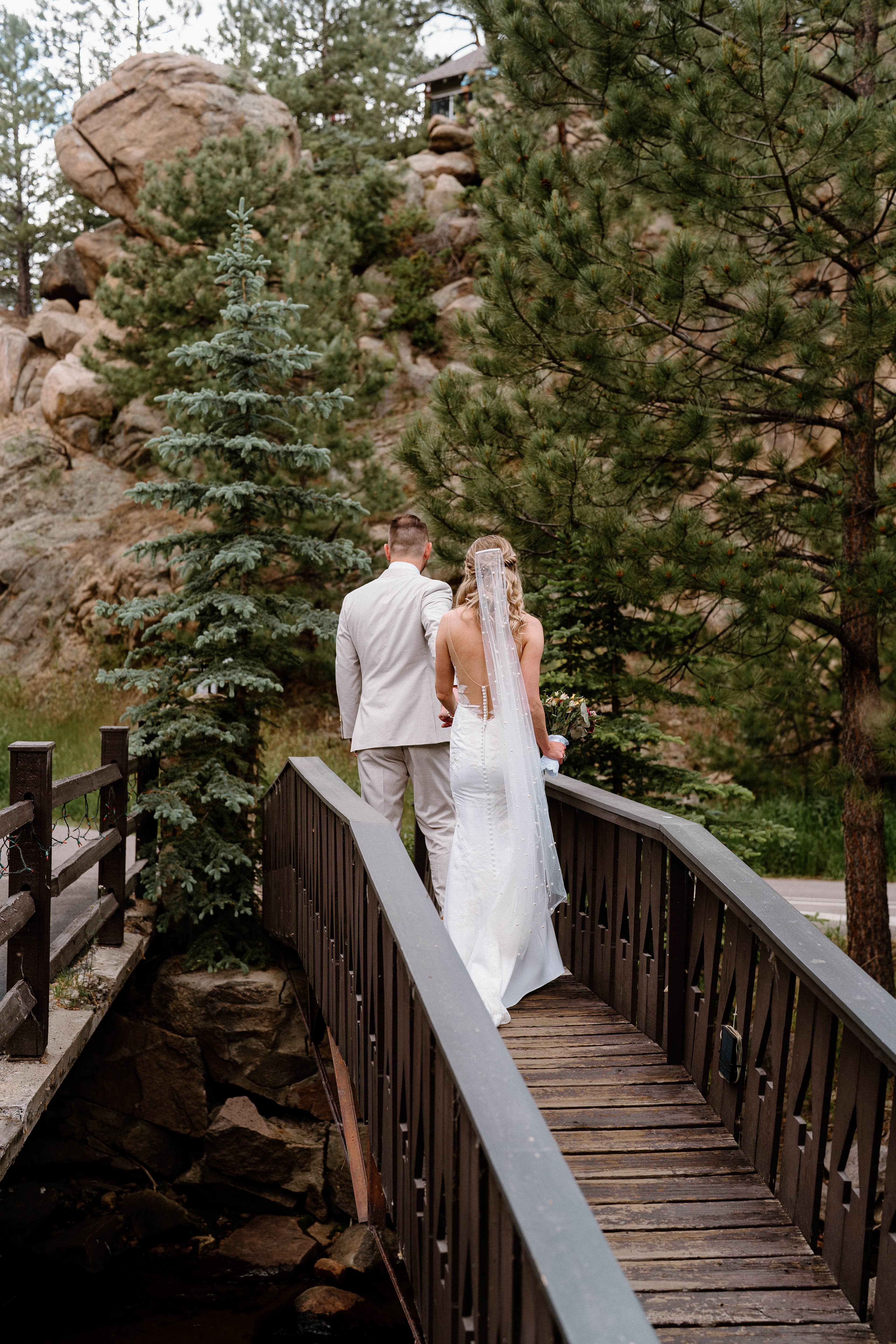 A bride and groom walk hand-in-hand across a bridge surrounded by trees and rocky terrain.