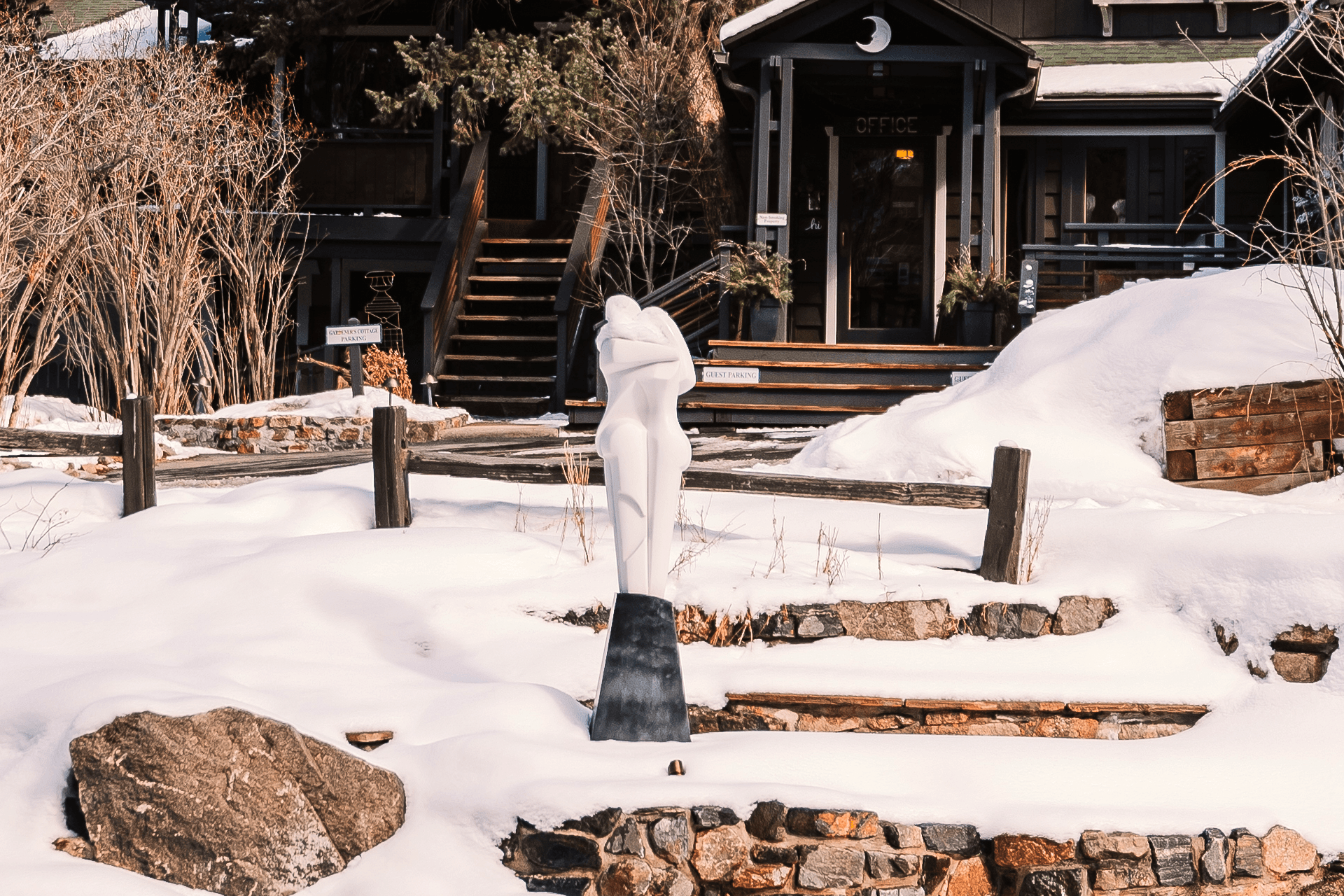 A white sculpture stands on a snowy landscape in front of a dark wooden building.