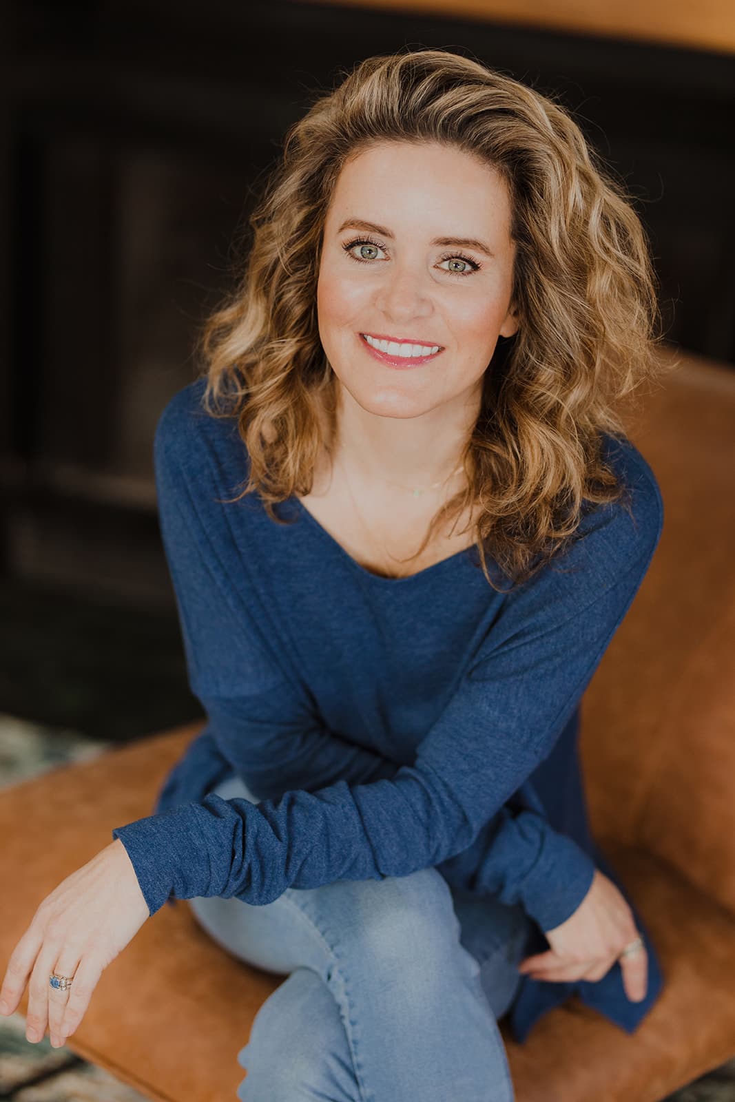 A smiling woman with curly hair sits casually on a brown leather couch, wearing a blue top and light jeans.