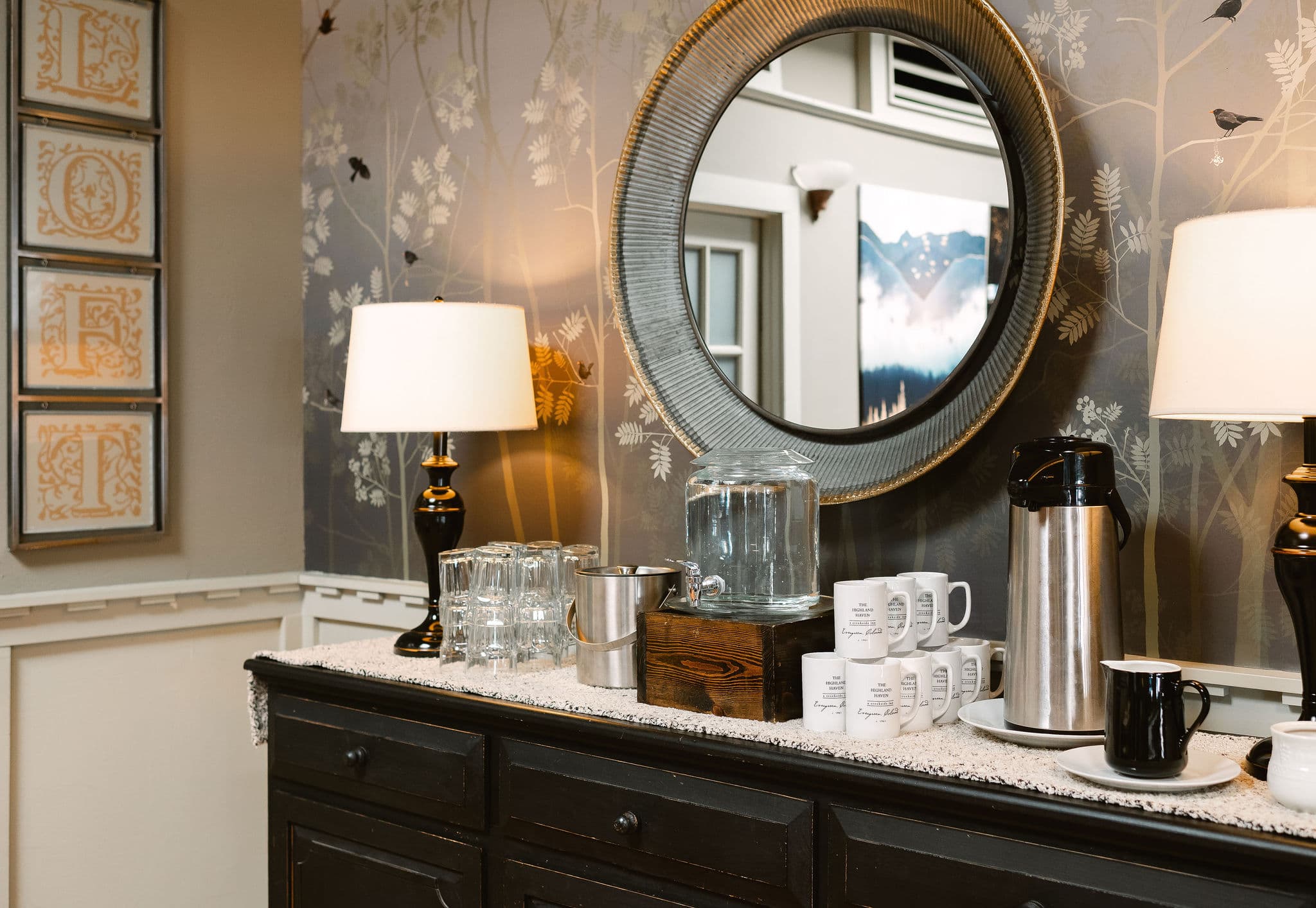 A decorated sideboard displays a water dispenser, coffee carafe, glasses, and mugs under a round mirror.