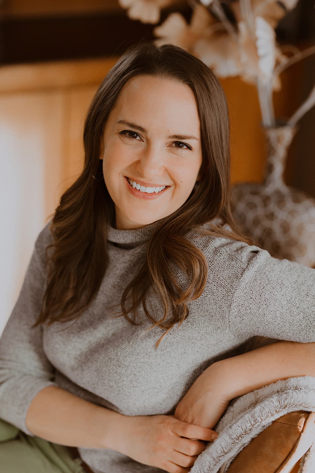 A woman with long brown hair smiles warmly while sitting comfortably in a cozy setting.