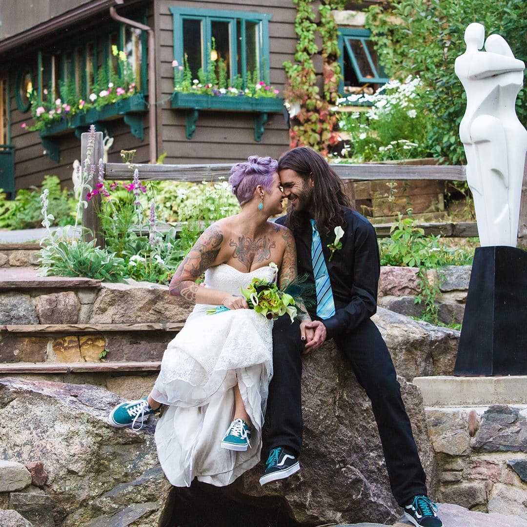 Tattooed bride with purple hair and groom with long hair and beard sit smiling on stone steps, wearing matching blue sneakers in a whimsical garden setting.