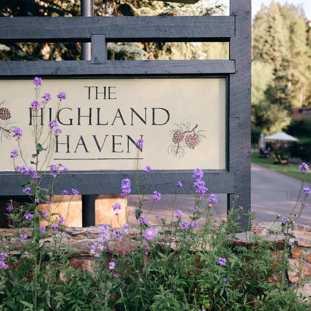 A rustic sign reading "The Highland Haven," surrounded by purple wildflowers, stands by a road lined with lush green trees, conveying a serene, welcoming atmosphere.