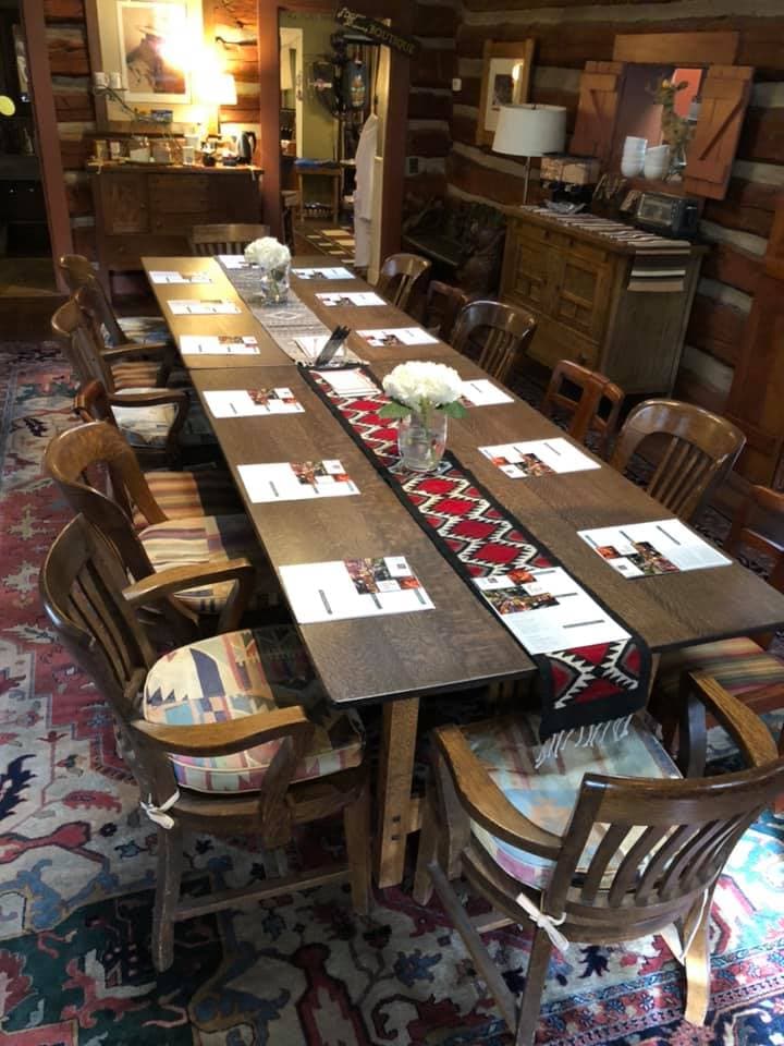 A long wooden dining table is set with place cards and flowers, surrounded by wooden chairs in a rustic interior.