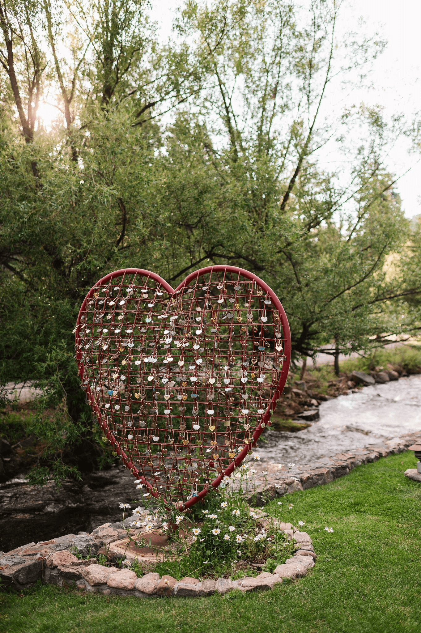 A large red heart-shaped structure adorned with padlocks stands near a flowing stream, surrounded by greenery and flowers.