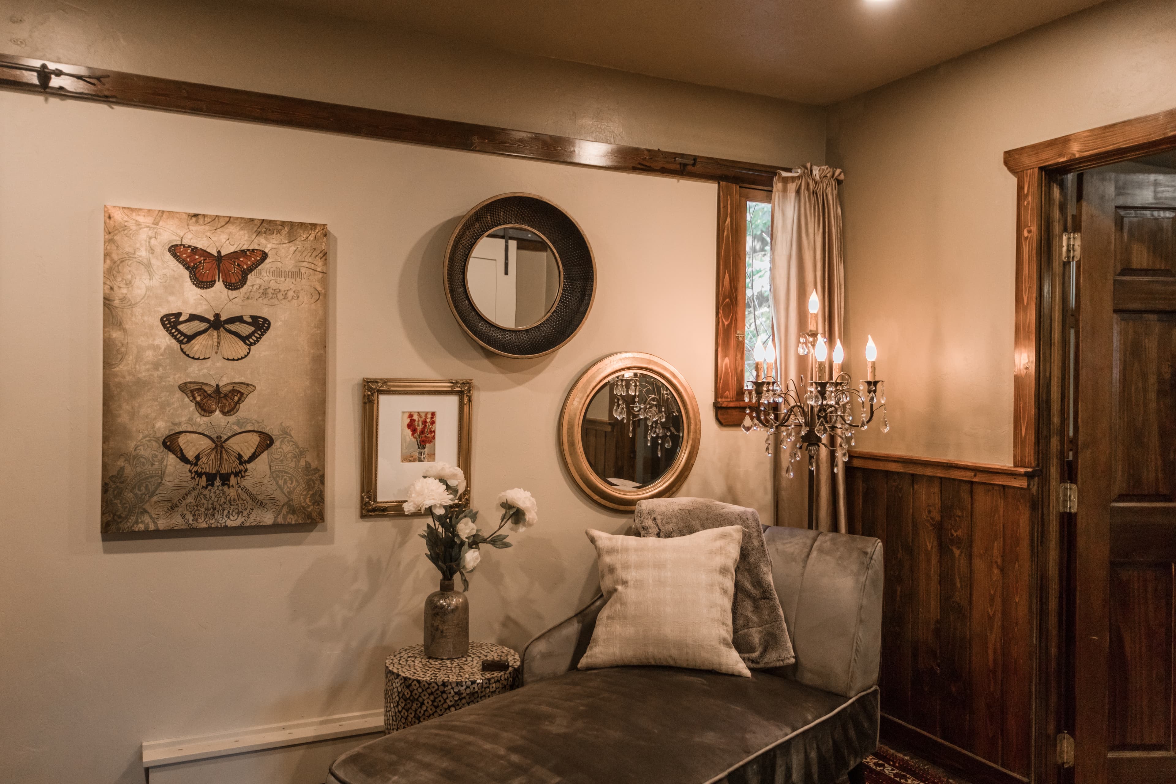 A cozy corner with a chaise lounge, decorative mirrors, a butterfly art piece, and a chandelier.
