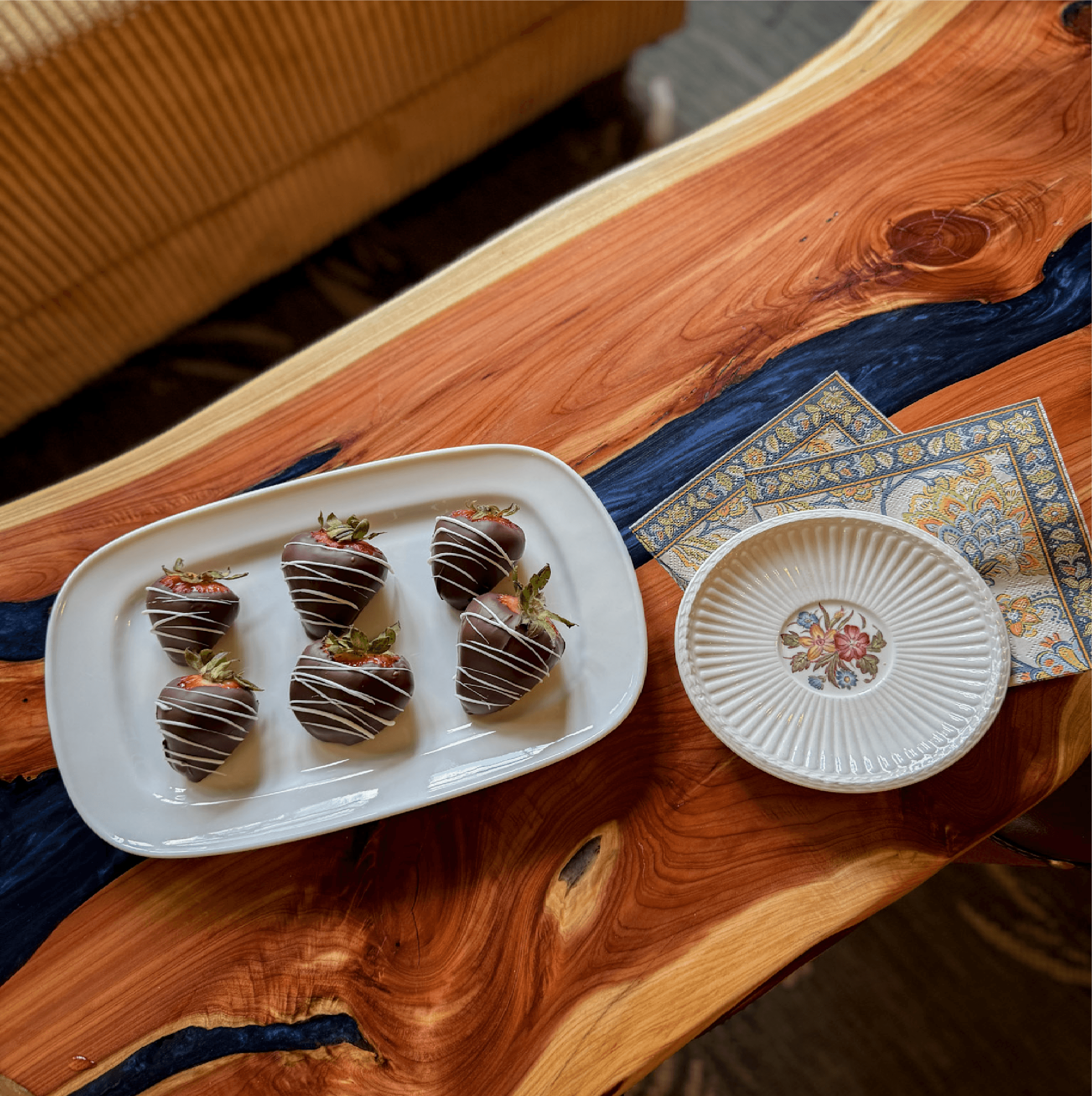 A platter of six chocolate-covered strawberries on a wooden table next to a decorative plate and napkins.