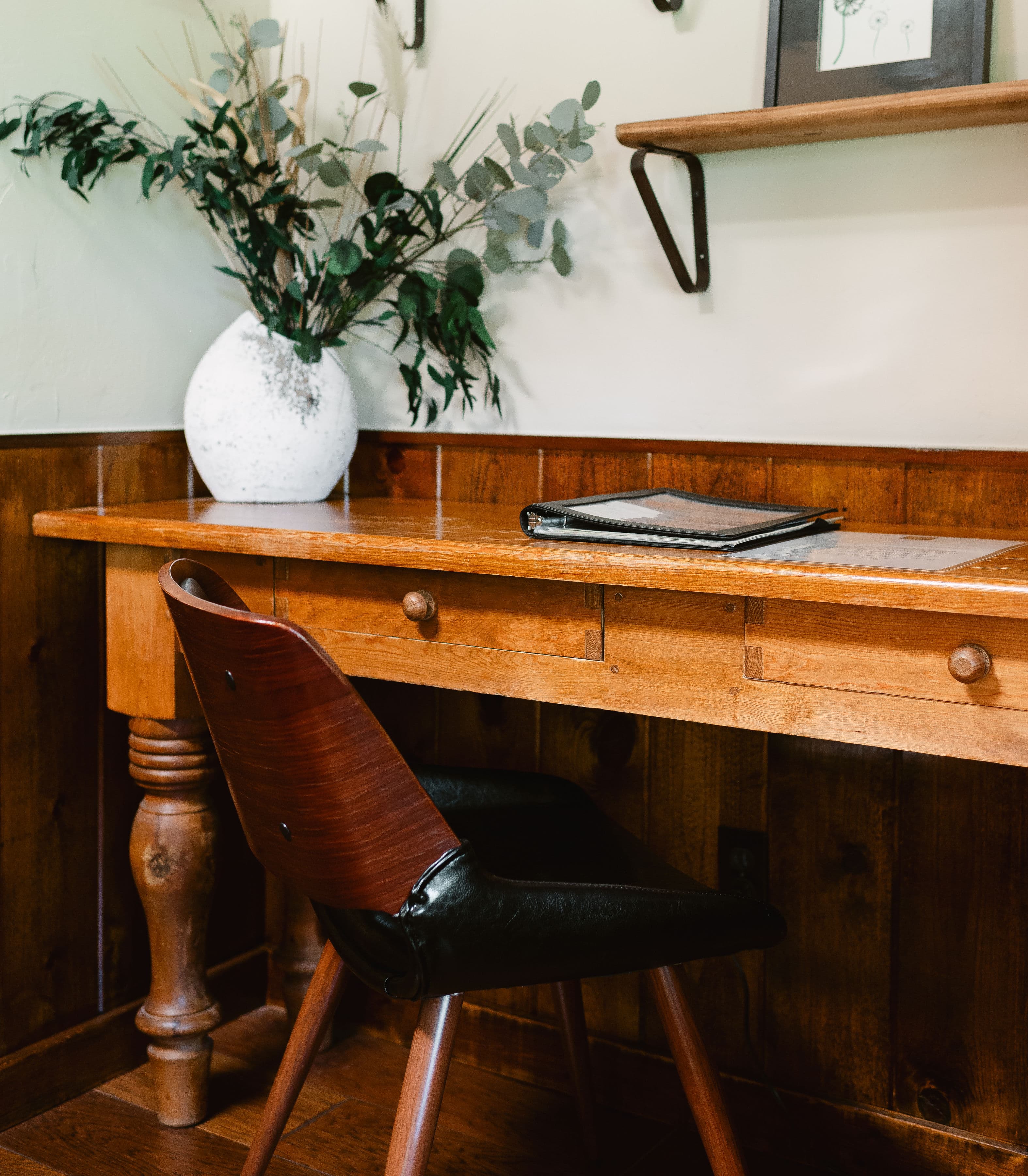 A wooden desk with a chair, a vase of greenery, and a black binder on top.