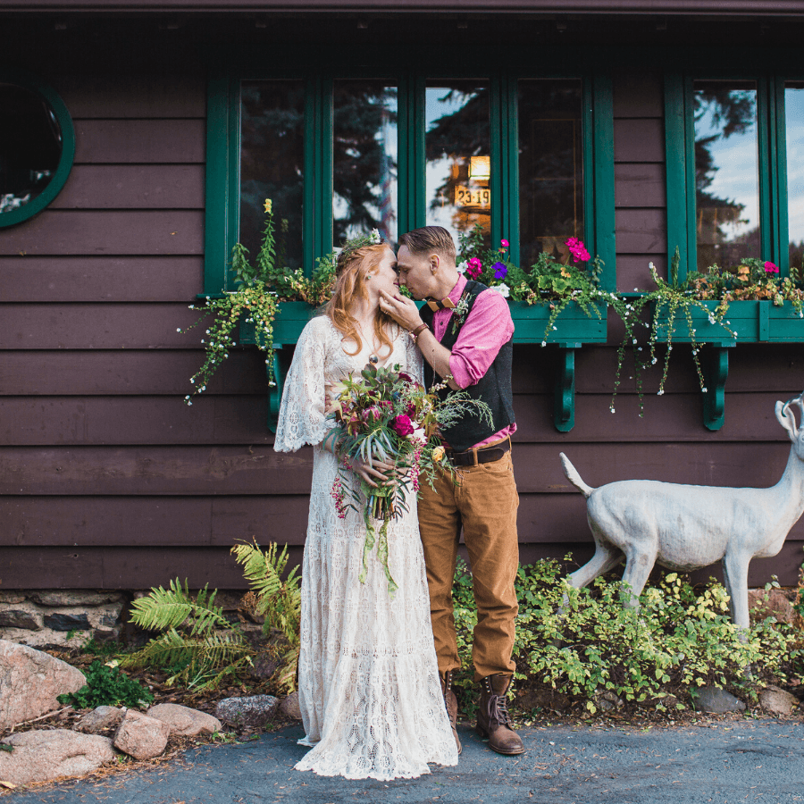 A couple leans in for a kiss in front of a rustic cabin adorned with flowers.