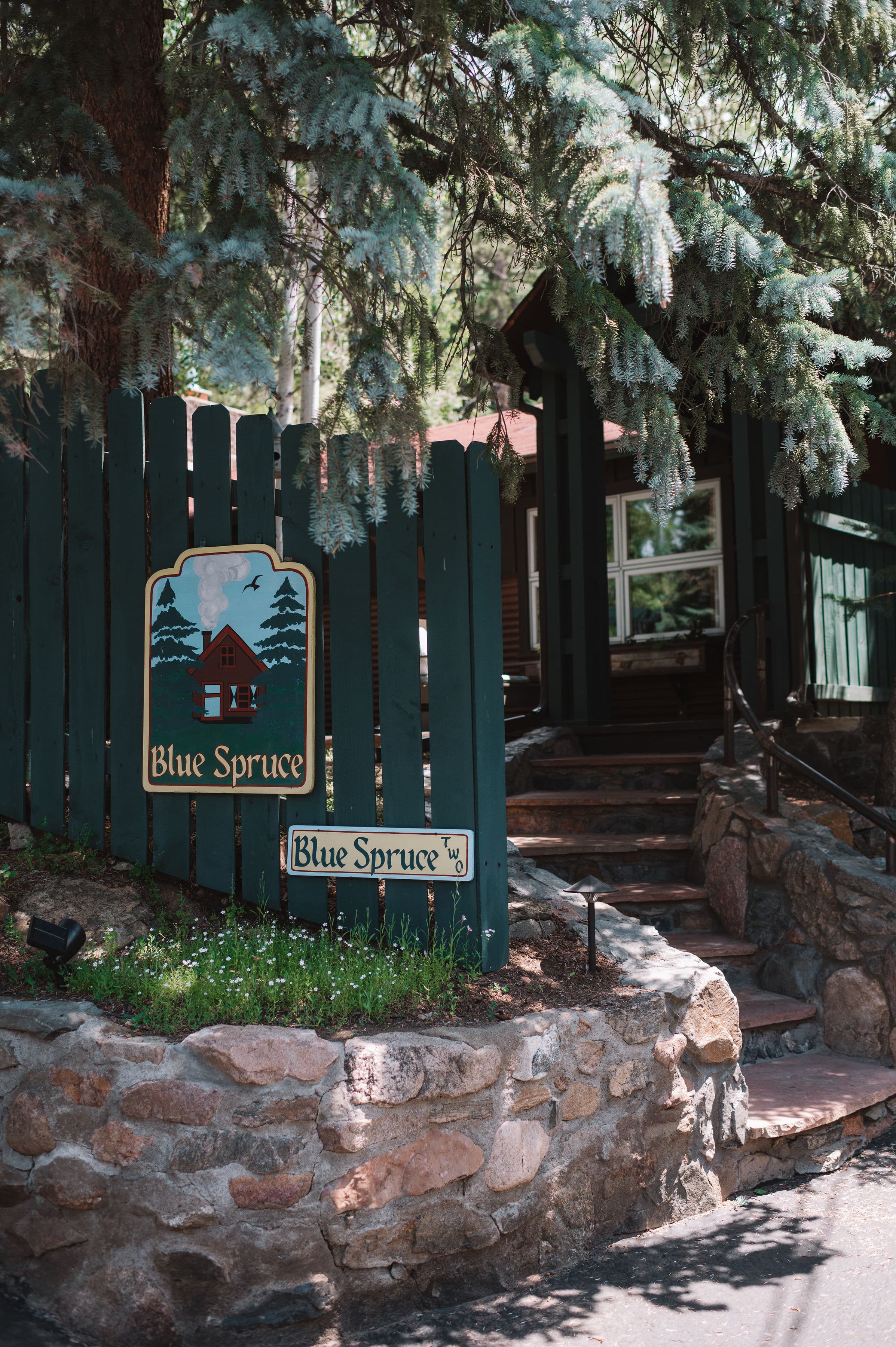 A green wooden fence with "Blue Spruce" signs and stone landscaping in a forested area.
