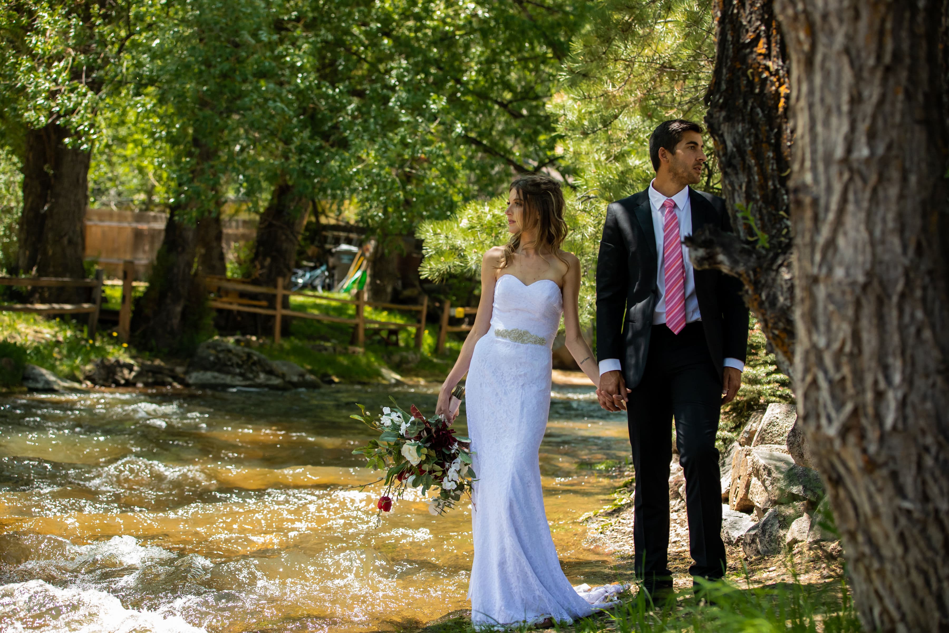 A bride and groom hold hands by a flowing stream, surrounded by lush greenery.
