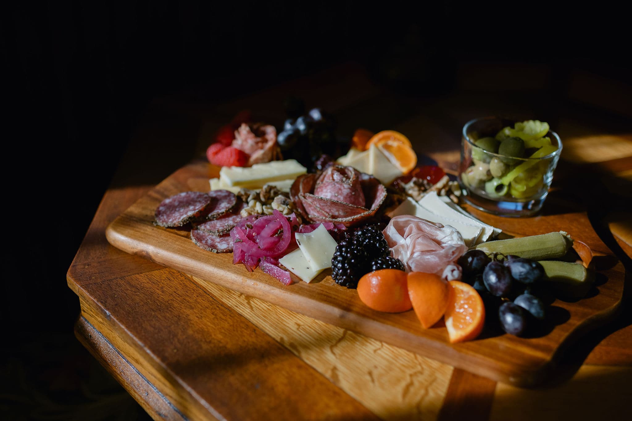 A wooden platter featuring a variety of meats, cheeses, fruits, and pickles.