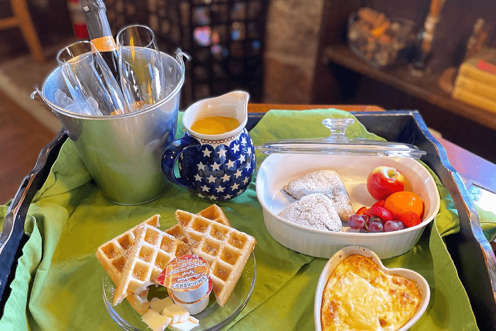Breakfast tray with heart-shaped cheese dish, waffles with butter and syrup, fruit, beignets, a blue star pitcher with juice, and ice bucket with glasses. Cozy setting by a fireplace.