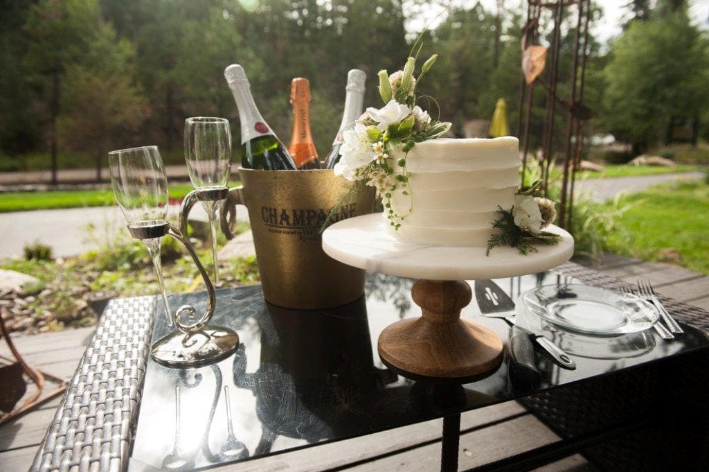 A white wedding cake with floral decorations is displayed on a wooden stand beside champagne and elegant glasses on a table.