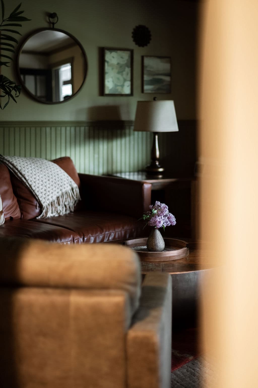 A cozy living room featuring a leather couch, a table with a vase of purple flowers, and framed artwork on the walls.