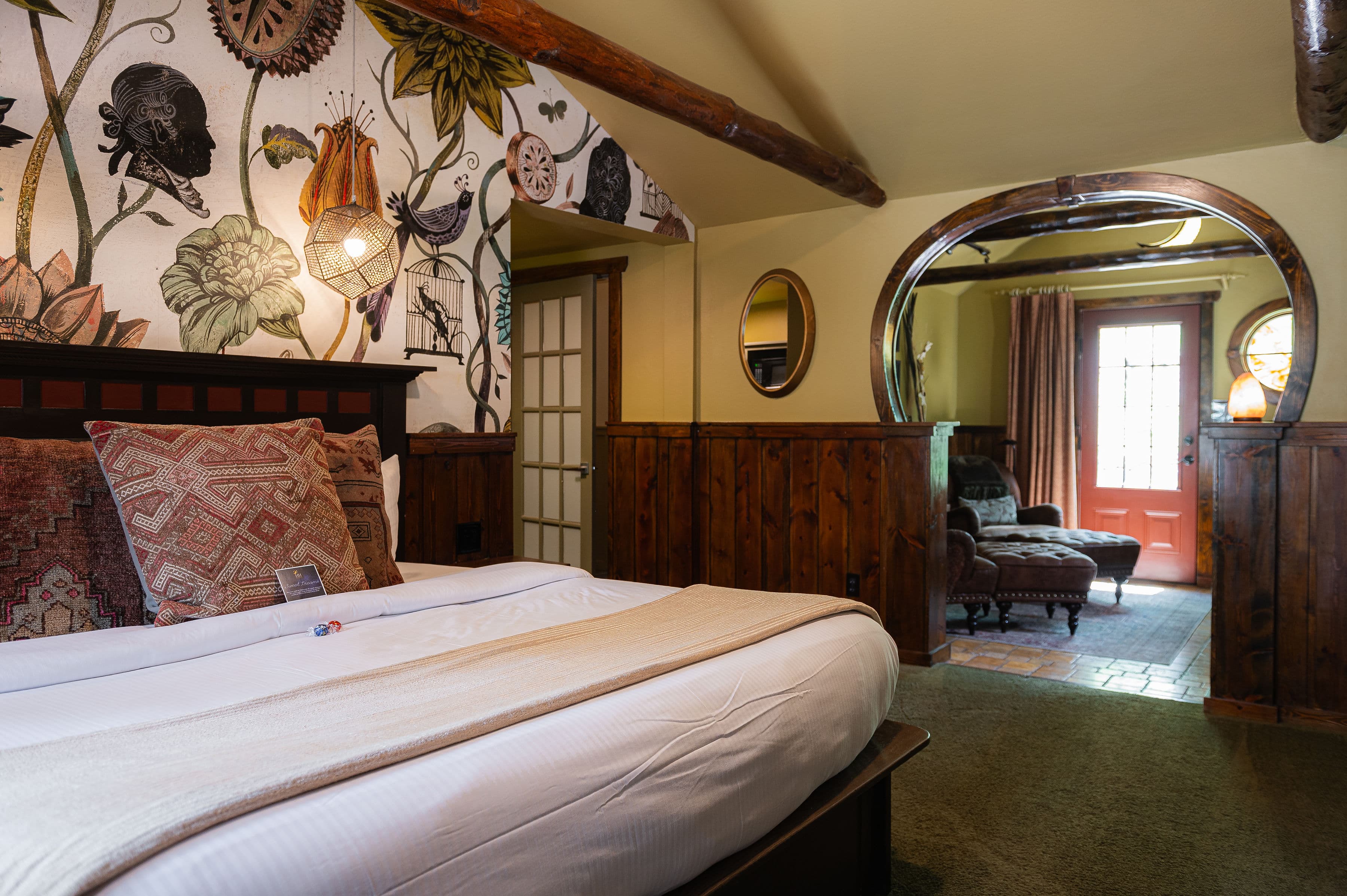A cozy bedroom featuring a decorative wall with floral and bird designs, a large bed, and a view of a door leading to another room.