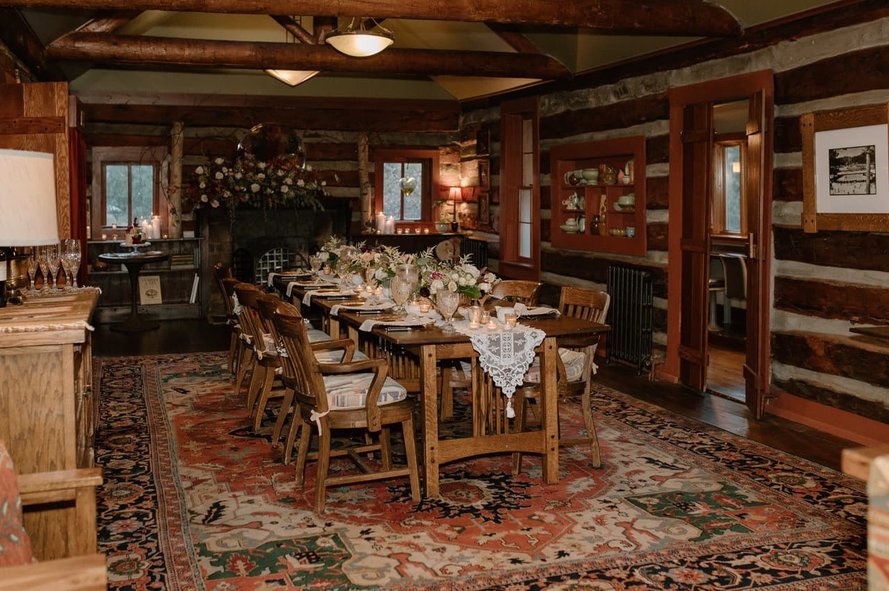 A rustic dining room featuring a long wooden table adorned with floral centerpieces, surrounded by wooden chairs, against a backdrop of log walls and a cozy fireplace.