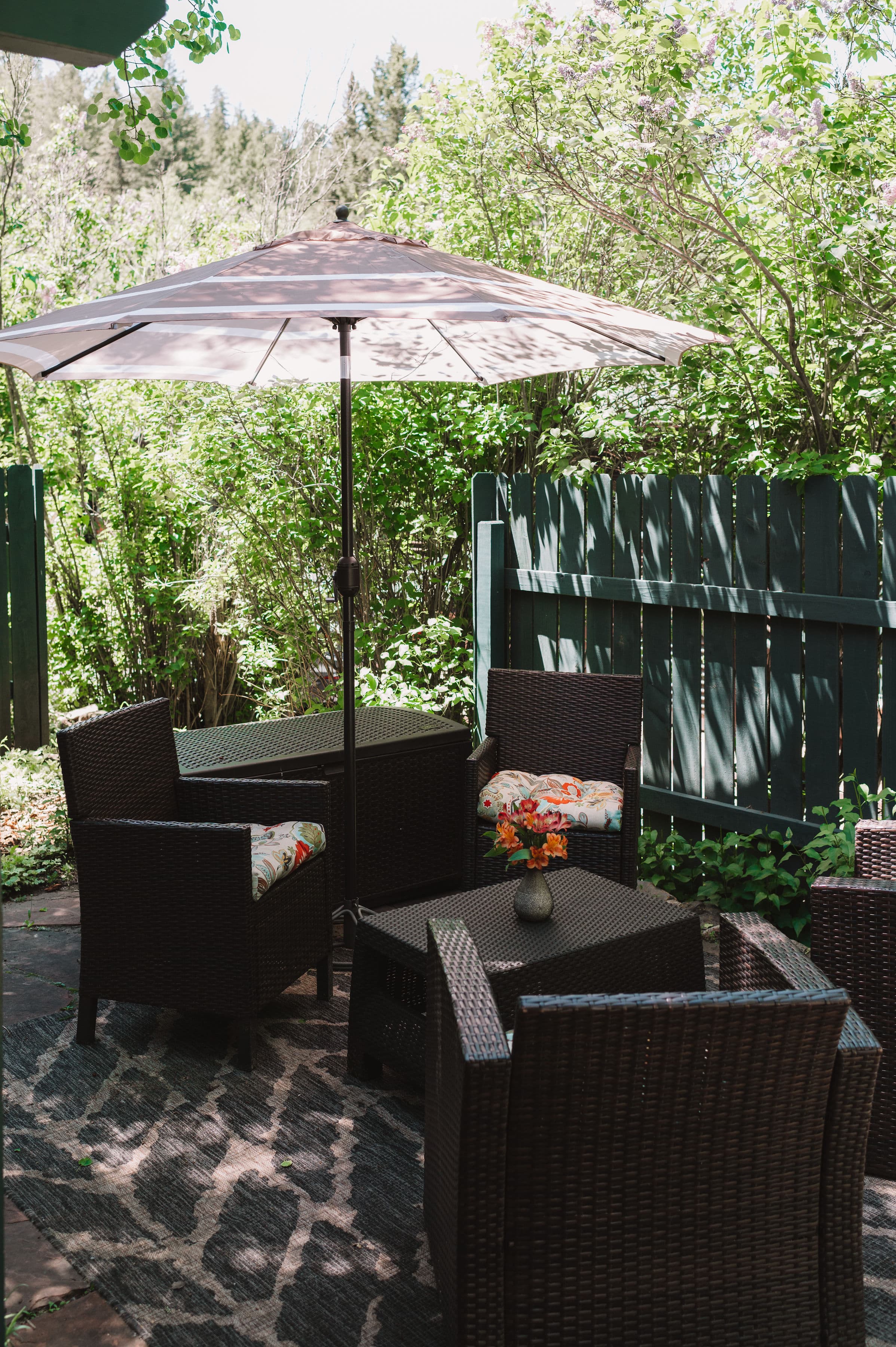 A cozy outdoor seating area with wicker chairs, a table, and a shaded umbrella surrounded by greenery.