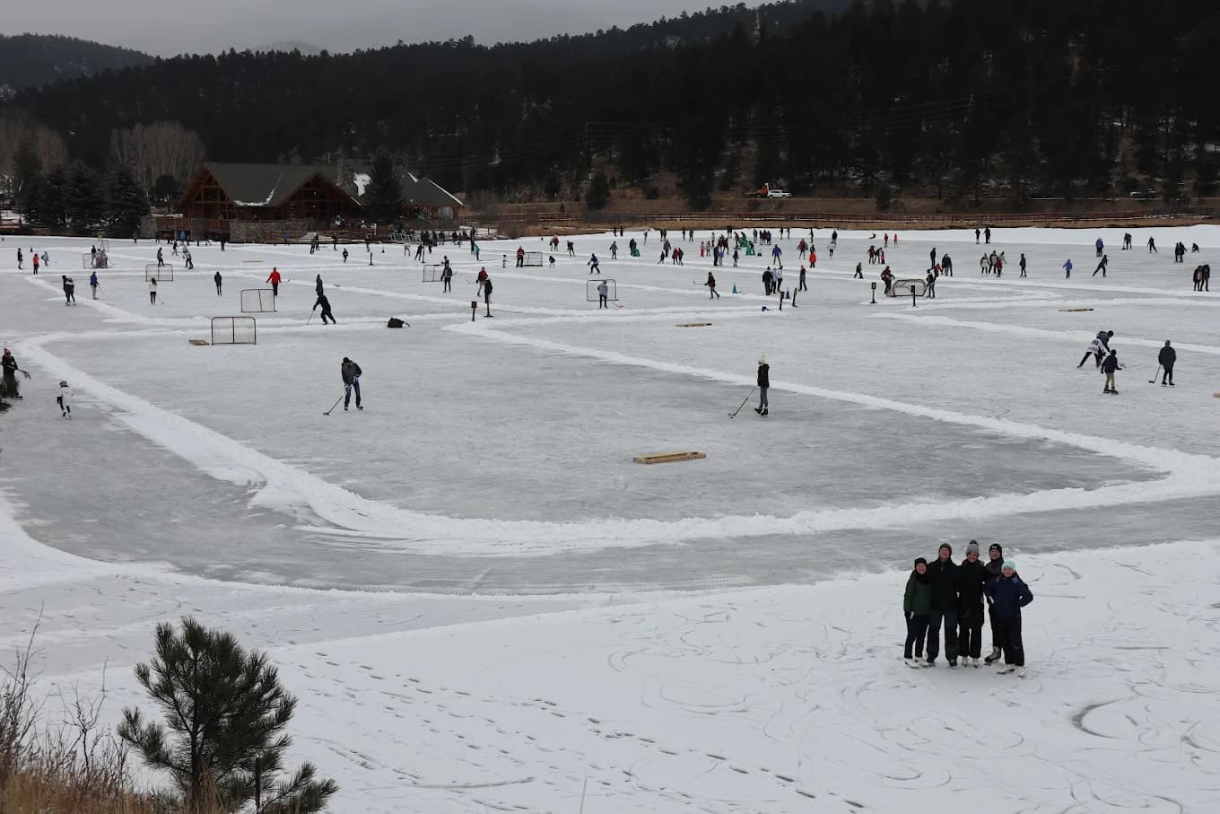 A large group of people ice skating and playing hockey on a frozen lake surrounded by snow-covered trees.