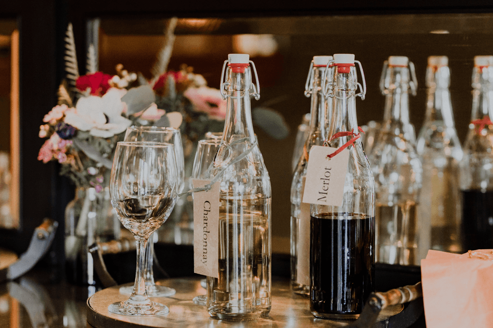 Bottles of Chardonnay and Merlot are displayed alongside a glass on a wooden table, with flowers in the background.