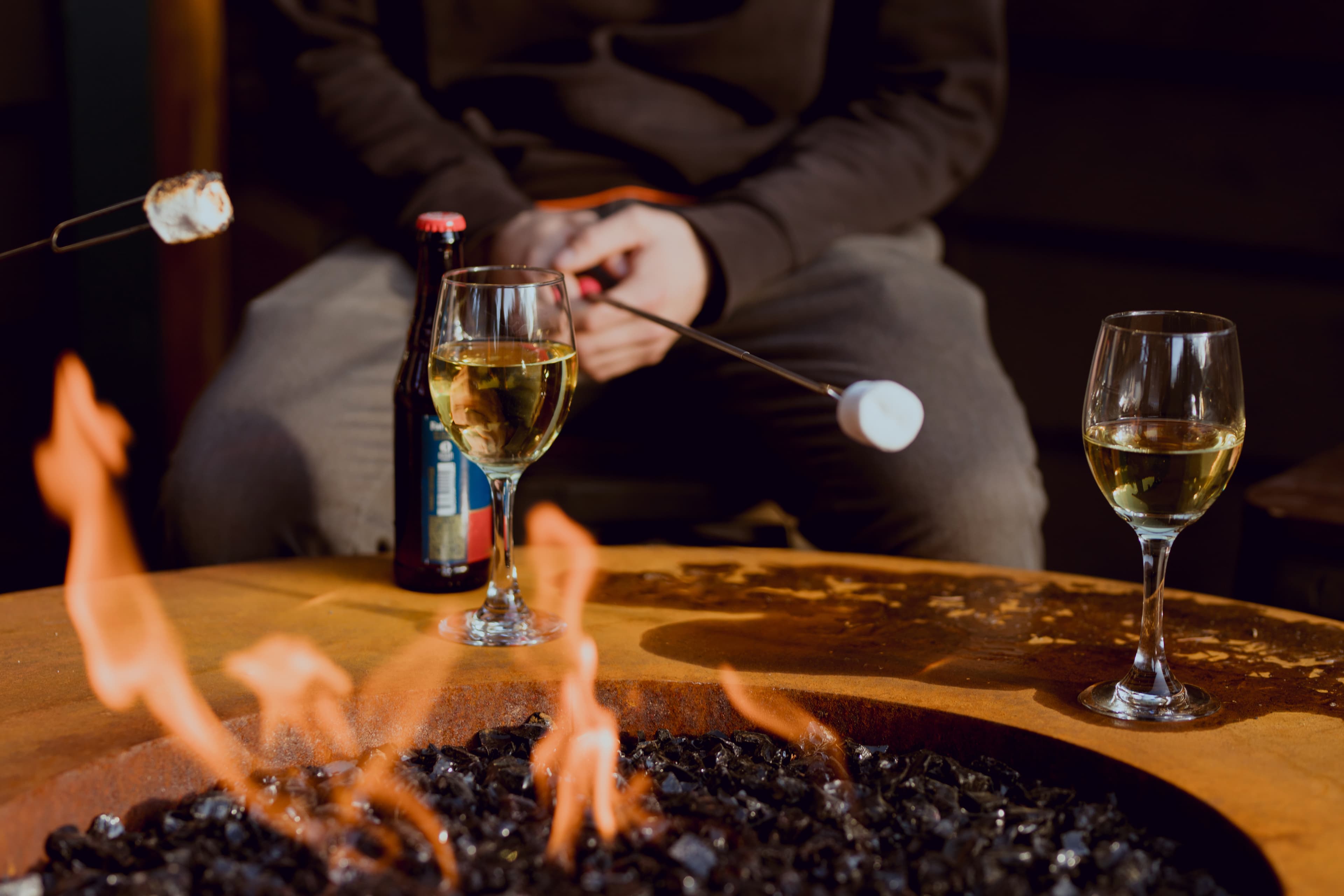 A person's hand holds a skewer with a marshmallow over a fire pit, with glasses of white wine and a beer bottle nearby.