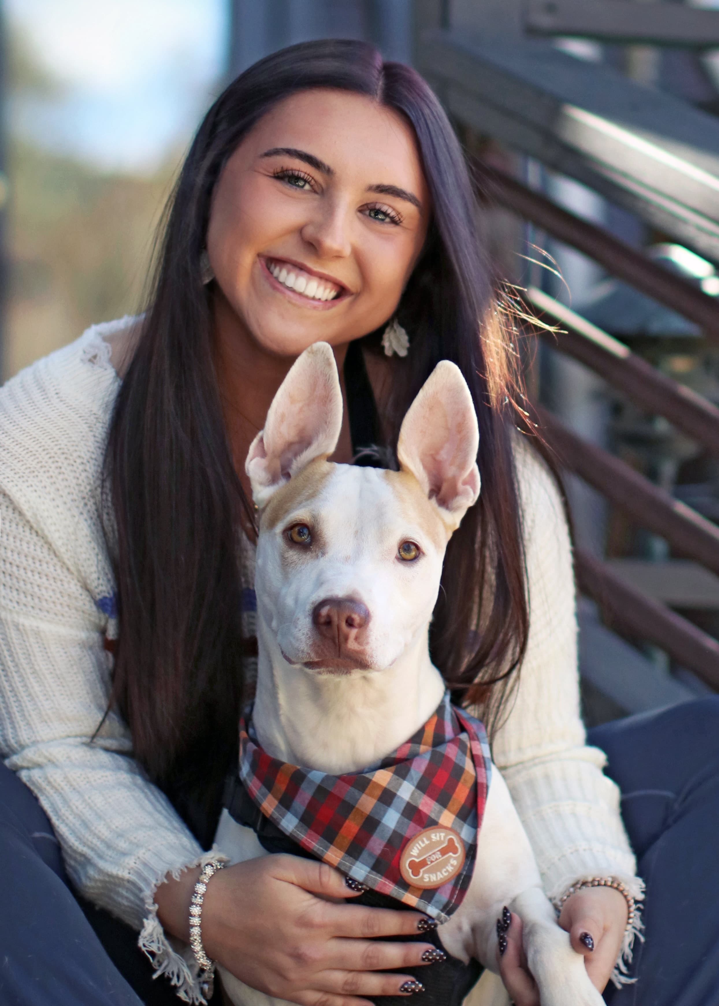 A smiling young woman sits on steps with a dog wearing a plaid bandana.