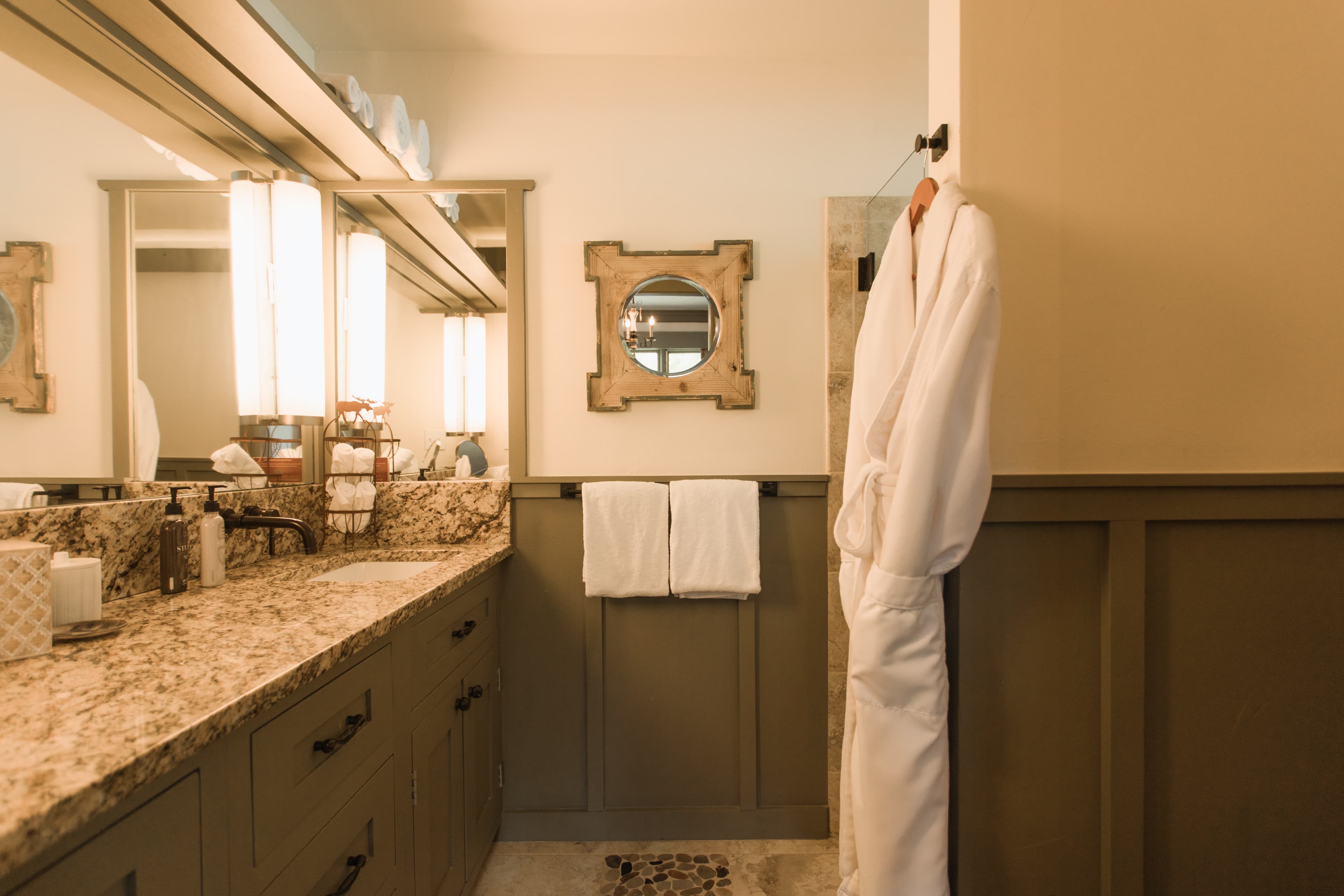 A modern bathroom featuring a granite countertop, two mirrors, hanging towels, and a white robe.