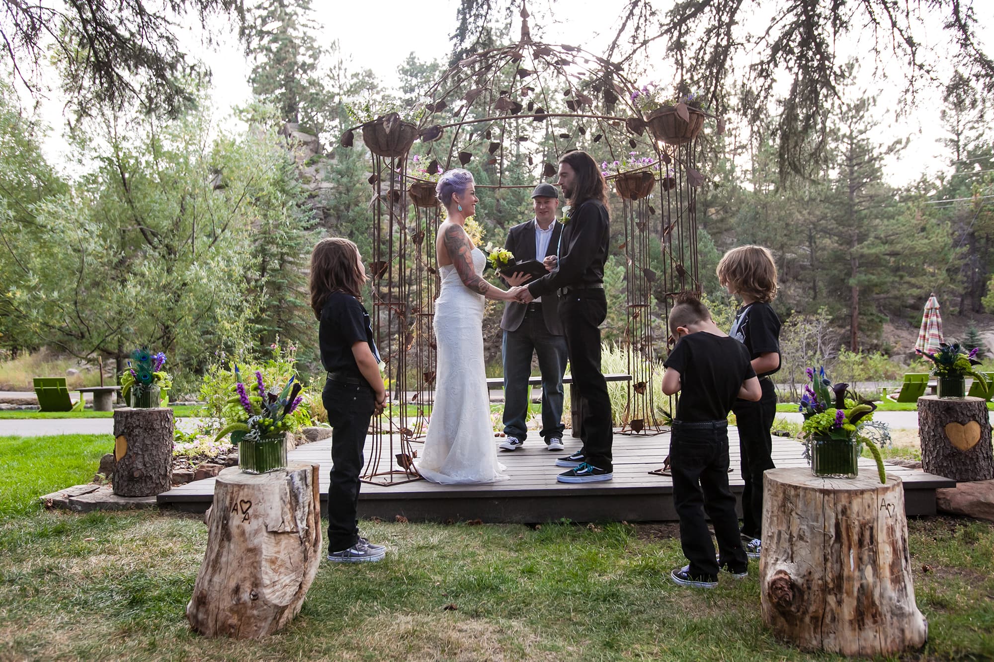 A couple stands together in an outdoor wedding ceremony surrounded by children and floral arrangements.