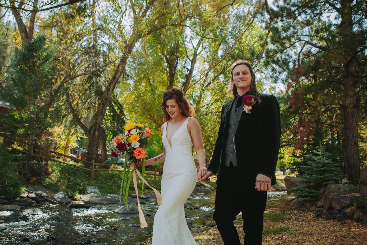 A bride and groom walk hand in hand by a sparkling stream surrounded by vibrant autumn foliage.
