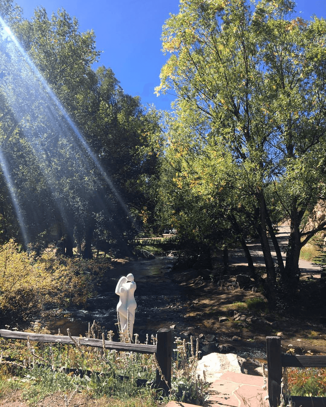 A white sculpture stands near a stream, surrounded by vibrant trees against a clear blue sky.