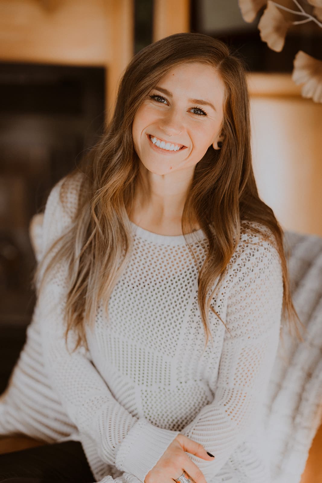 A smiling woman with long hair sits casually in a cozy setting.