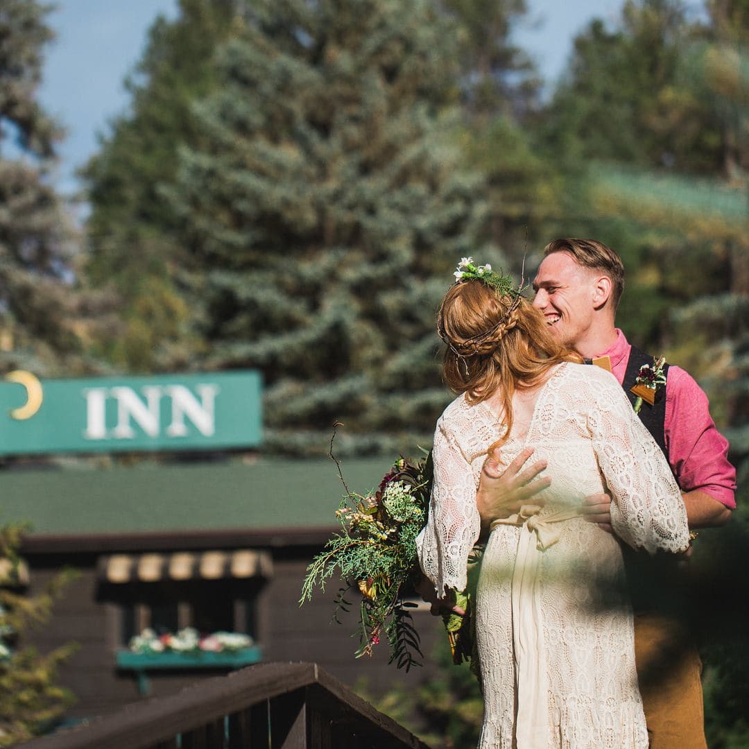 A bride and groom embrace joyfully outdoors, surrounded by trees and a sign for an inn in the background.