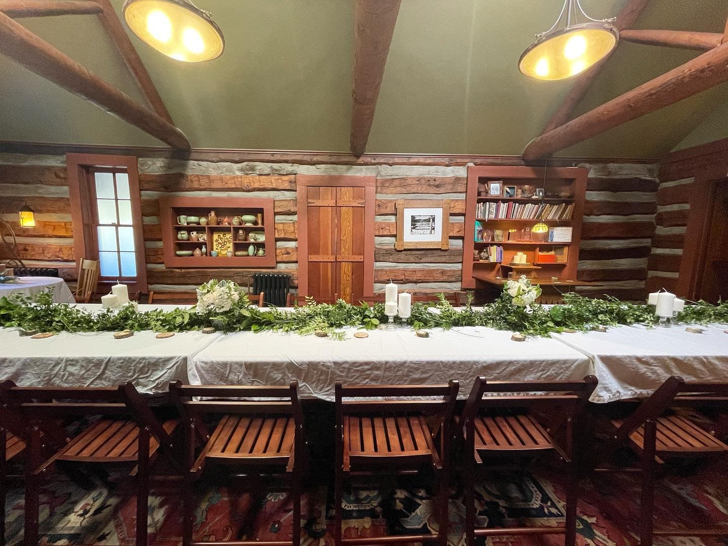 A rustic dining room with a long table adorned with greenery and candles, surrounded by wooden chairs and log walls.