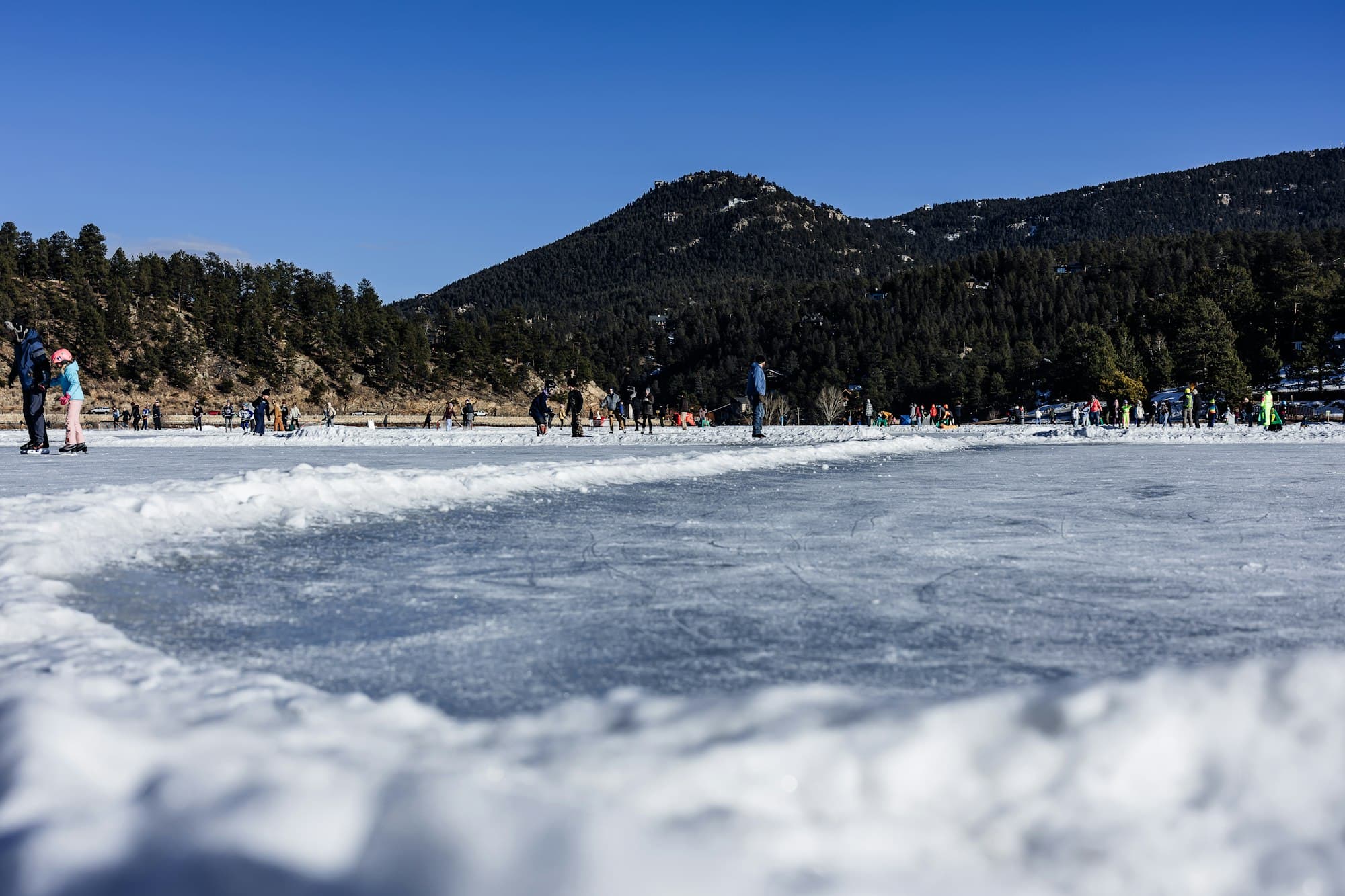 A frozen lake with people ice skating and a mountainous backdrop under a clear blue sky.
