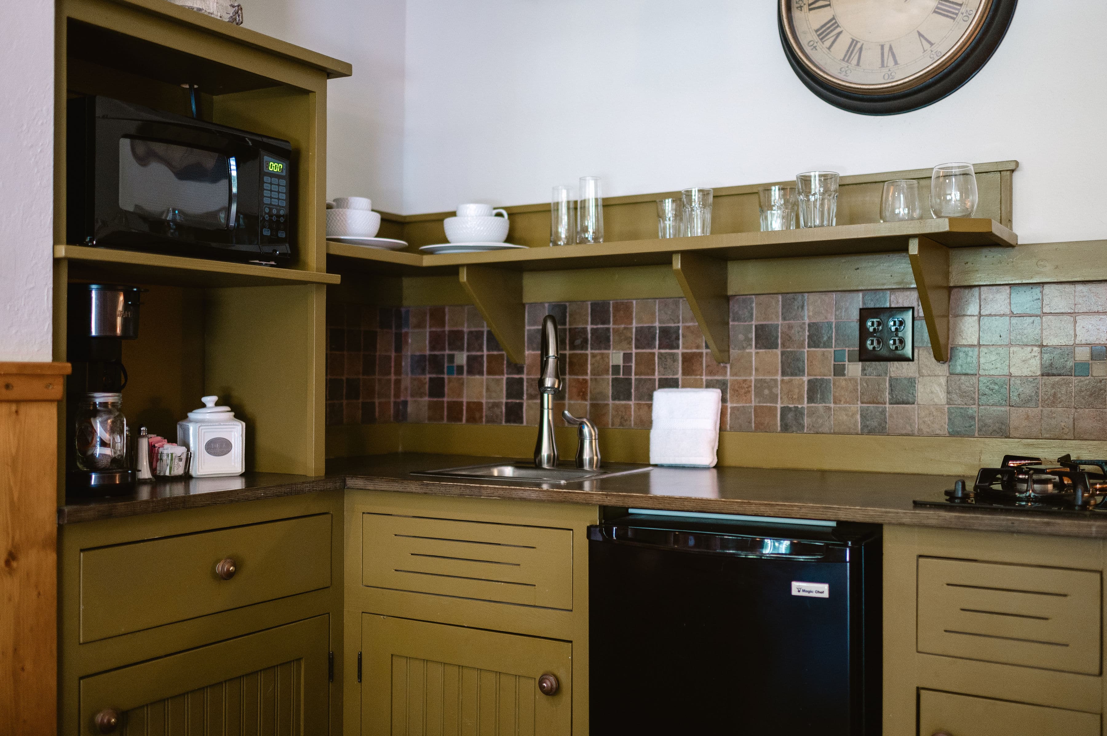 A compact kitchen with green cabinetry, a microwave, coffee maker, and a sink, set against a tiled backsplash.