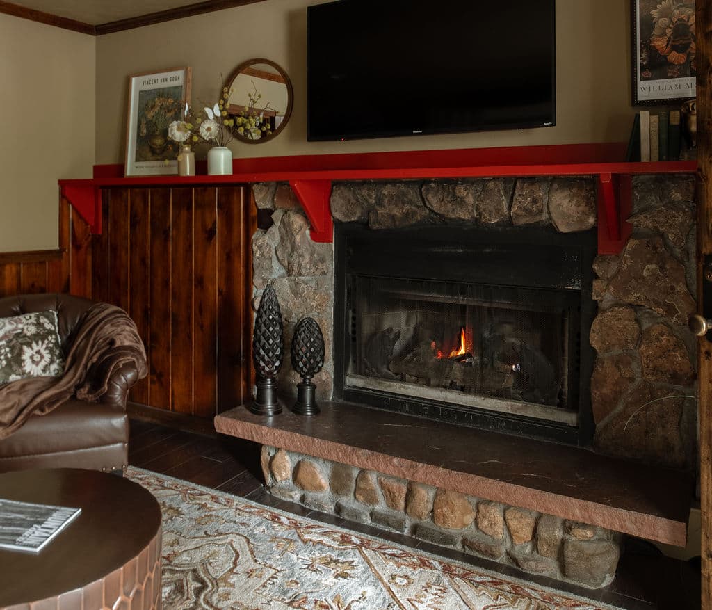 A cozy living room featuring a stone fireplace with a flickering flame, a brown leather couch, and a red mantel.