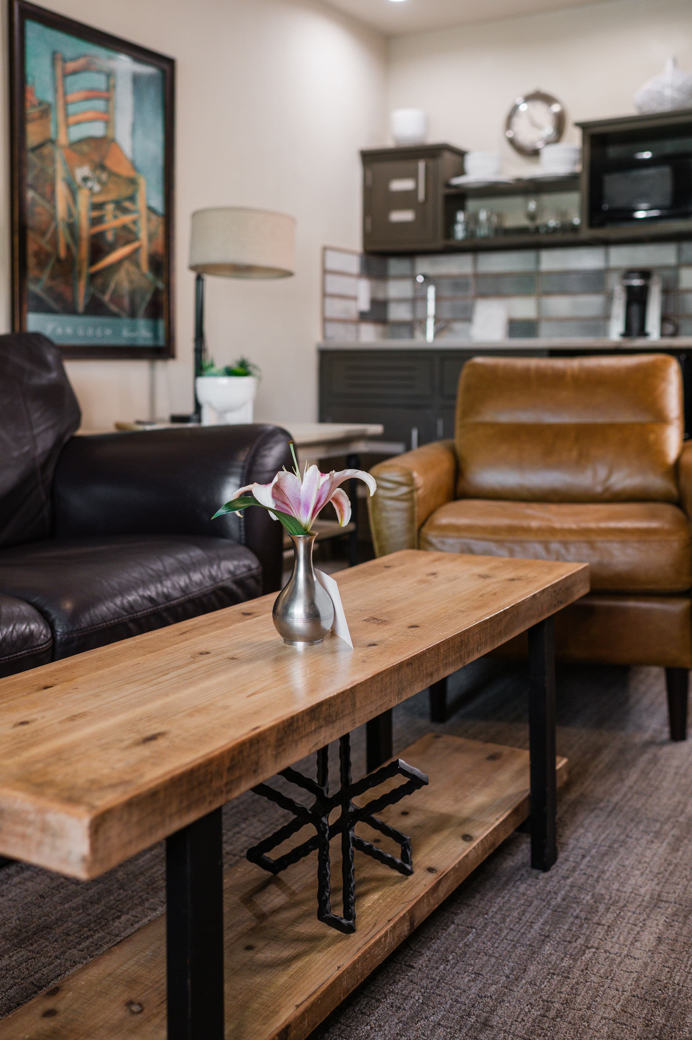 A cozy sitting area featuring a wooden coffee table with a vase of flowers, flanked by leather chairs and modern kitchen cabinets in the background.