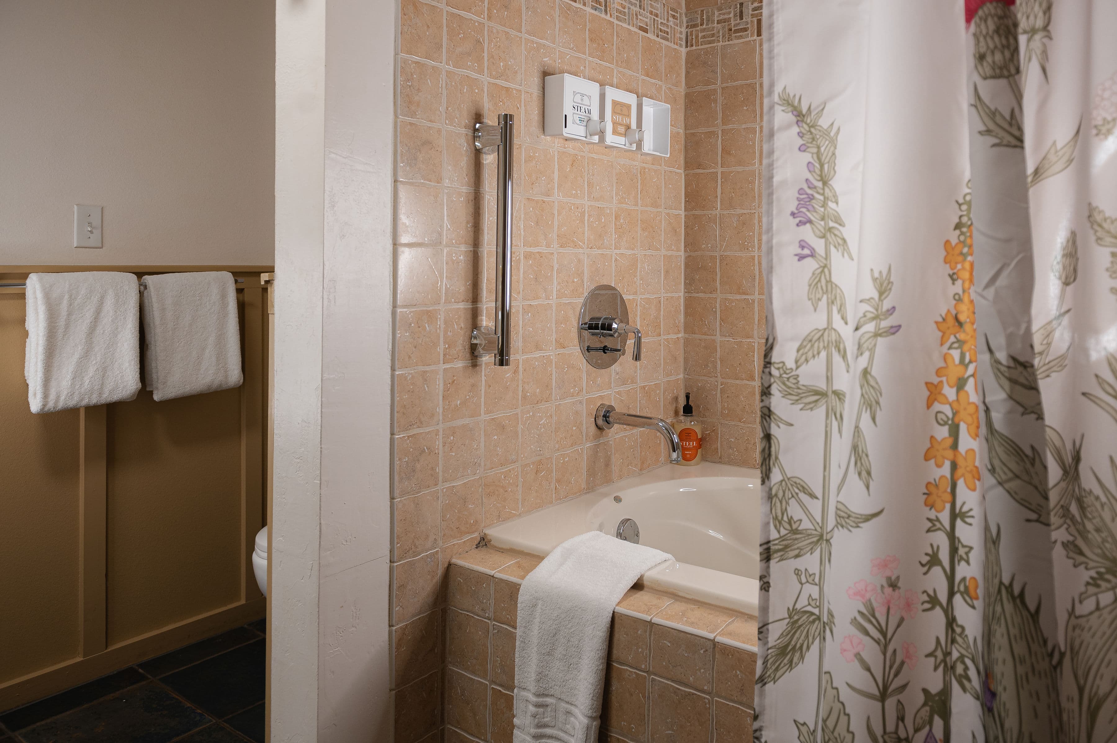 A bathroom featuring a tiled bathtub, a shower curtain with floral designs, and neatly hung white towels.