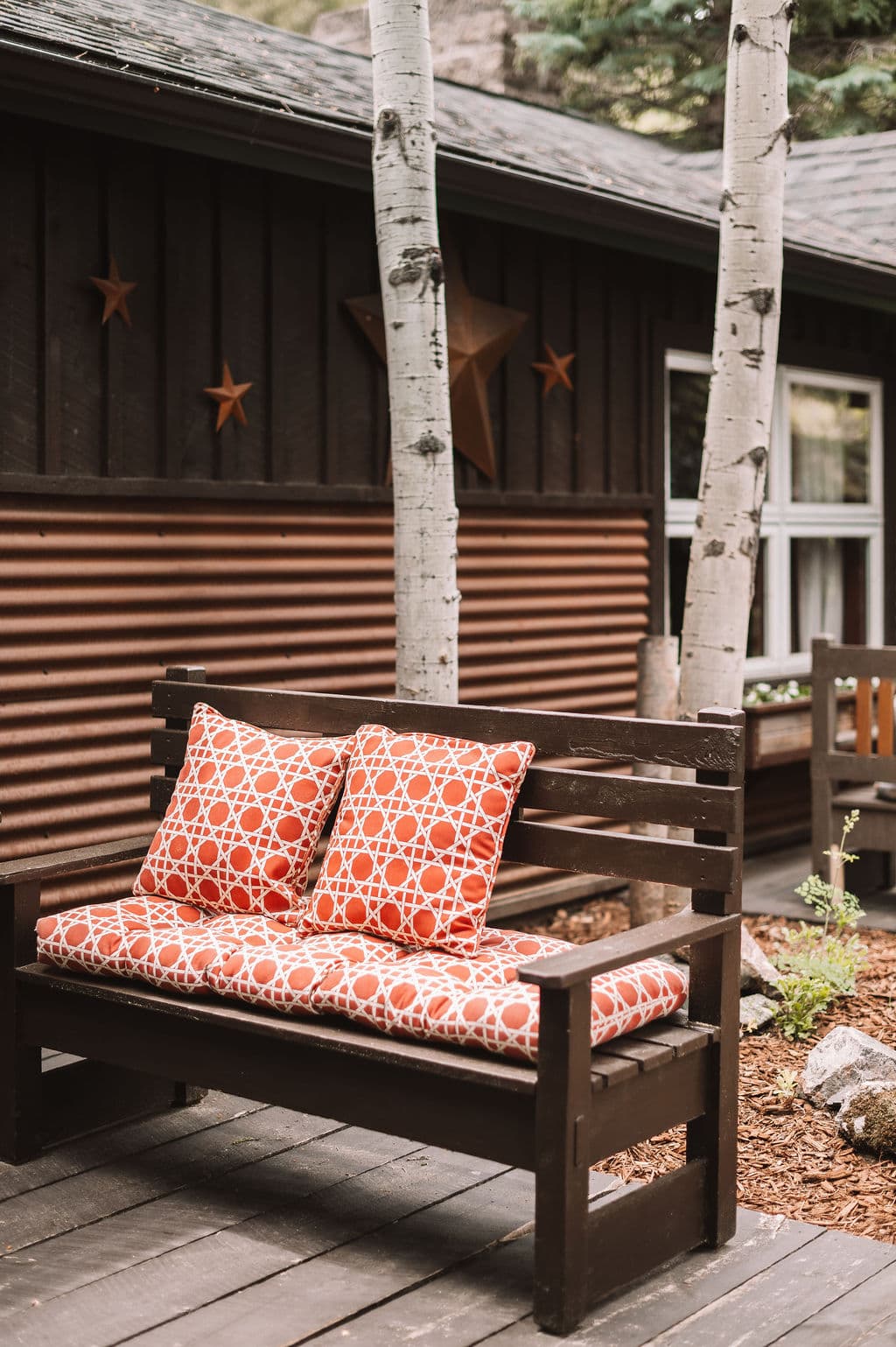 A wooden bench with orange patterned cushions is positioned beside a cabin and aspen trees.
