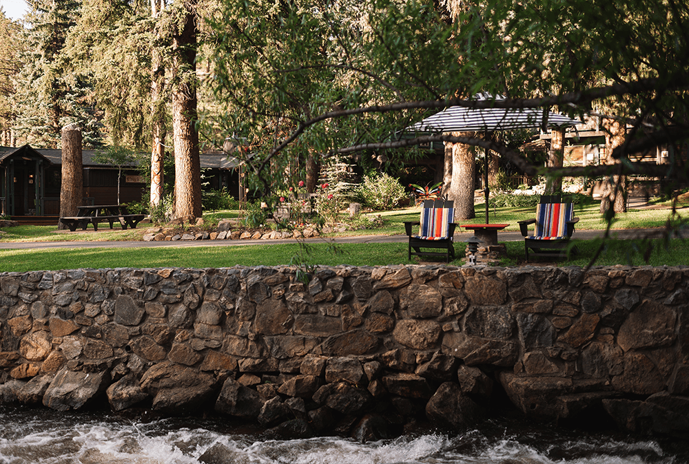 A serene forest scene with two colorful lounge chairs and an umbrella on a lush lawn behind a stone wall. A gentle river flows in the foreground.