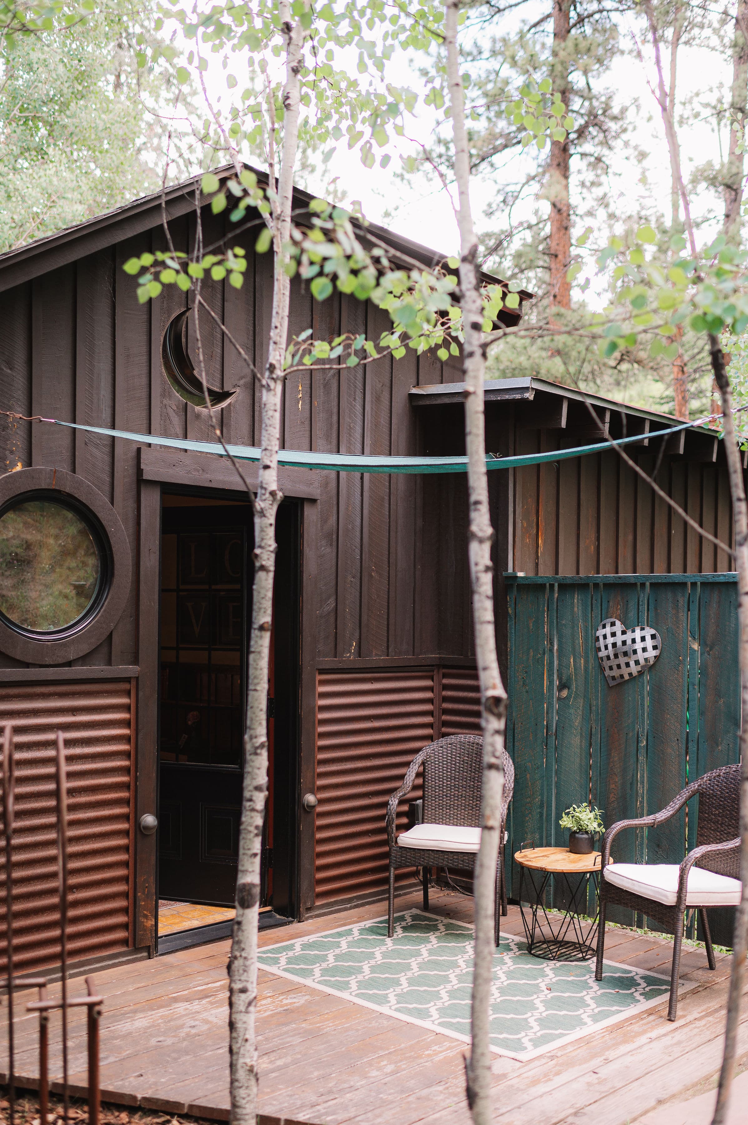 A cozy cabin entrance surrounded by trees, featuring two chairs and a small table on the porch.