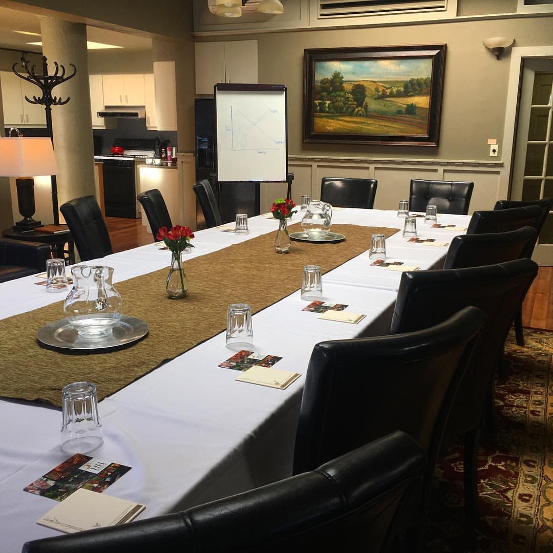 A conference room set with a long table, black chairs, and floral centerpieces, featuring a whiteboard and kitchen area in the background.