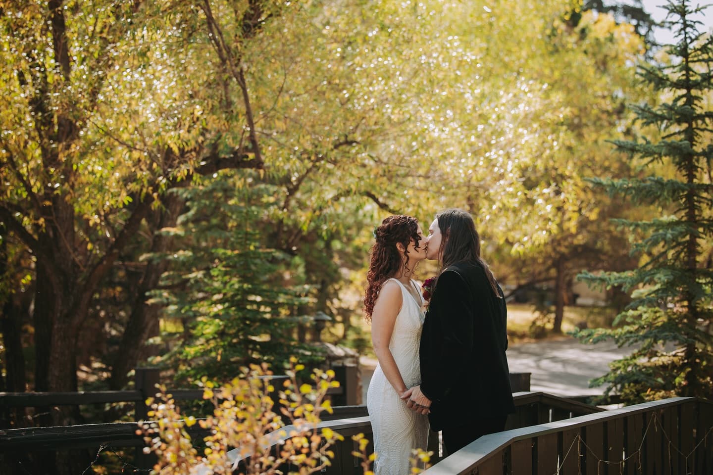 A couple shares a kiss on a bridge surrounded by autumn foliage.