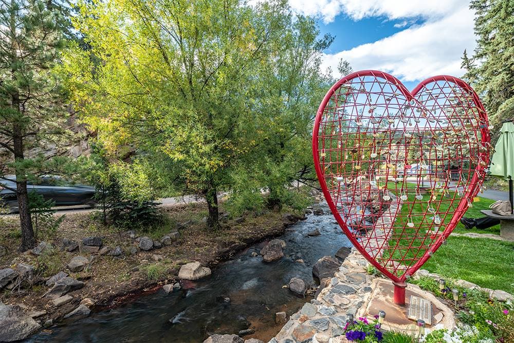 A large red heart sculpture stands by a flowing stream, surrounded by trees and greenery.